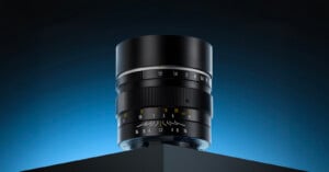 A sleek black camera lens with yellow and white markings is displayed upright on the edge of a black geometric surface, against a blue gradient background.