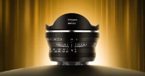 A 7Artisans 6mm f/2.0 camera lens is displayed upright on a dark surface, with a glowing golden background and vertically streaked light, highlighting its wide, curved front element and detailed focus markings.