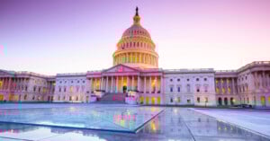 The United States Capitol building at sunrise, with its dome centered, glowing lights inside, and a pink-purple sky. The wet pavement in the foreground reflects the architecture.