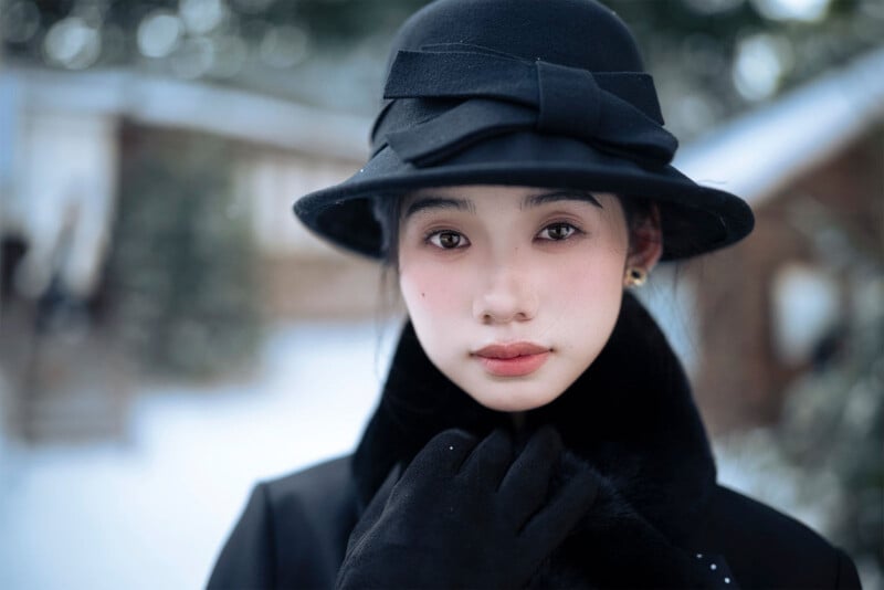A young woman wearing a black hat, gloves, and coat stands outdoors in a snowy setting, gazing directly at the camera with a calm expression. Blurred buildings and trees are visible in the background.