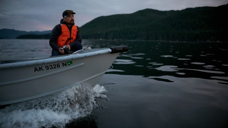 A man wearing an orange life jacket sits in a small boat moving across a calm lake, with forested hills in the background under a cloudy sky. The boat has the registration number "AK 9326 N.