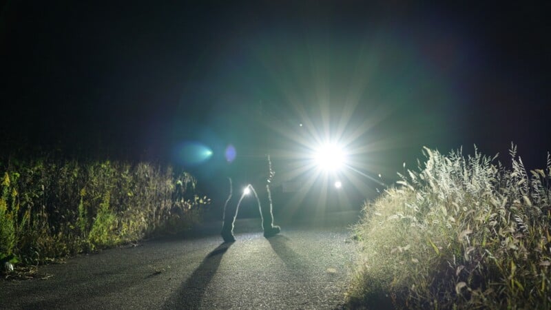 A person stands on a dark road at night, silhouetted by bright headlights from a vehicle facing them; tall grass lines both sides of the road, illuminated by the light.