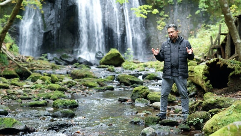A man in a black jacket stands on mossy rocks in a forest stream, gesturing with his hands. Behind him, a waterfall cascades over dark rocks surrounded by green trees and foliage.