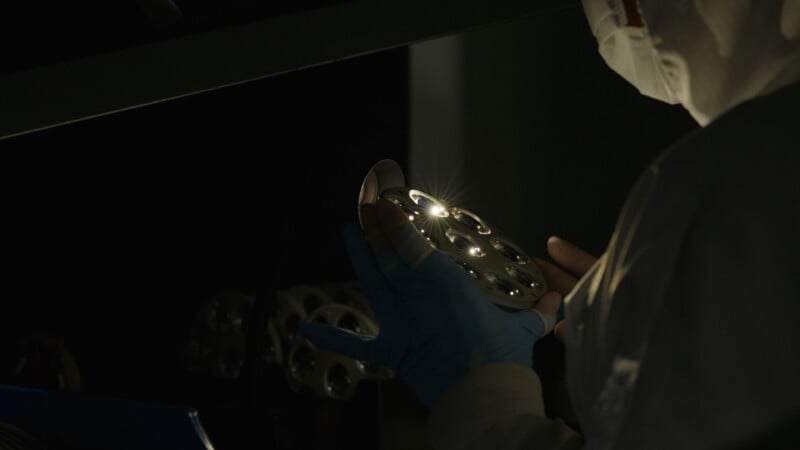 A person in protective clothing and blue gloves inspects a shiny, circular metallic object with holes, illuminated by a focused light in a dark environment.