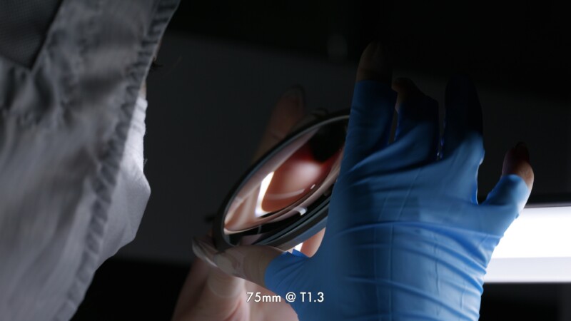 A person wearing a blue glove and protective clothing holds a camera lens element up to a bright light for inspection. Text at the bottom reads, "75mm @ T1.3.