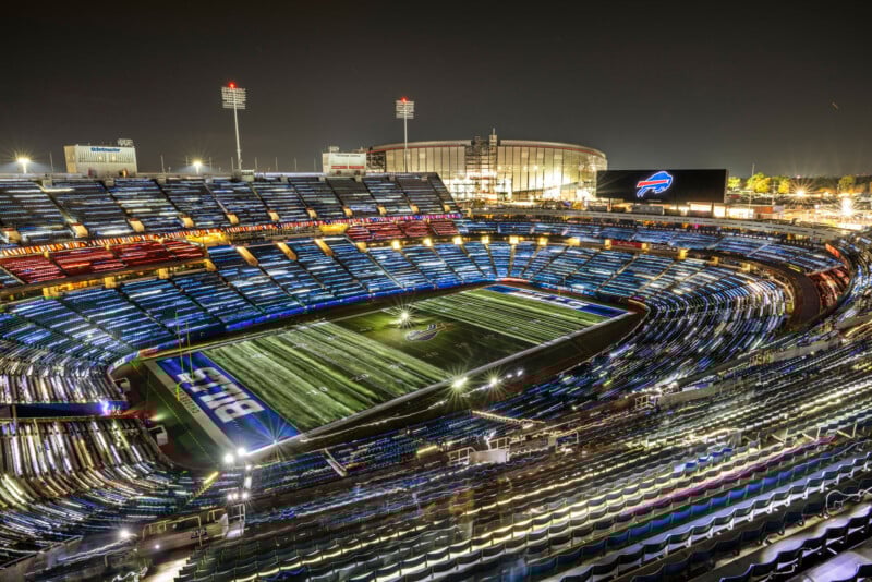 A brightly lit football stadium at night, with empty stands and a Buffalo Bills logo on the field and scoreboard. The arena glows under stadium lights and the night sky.