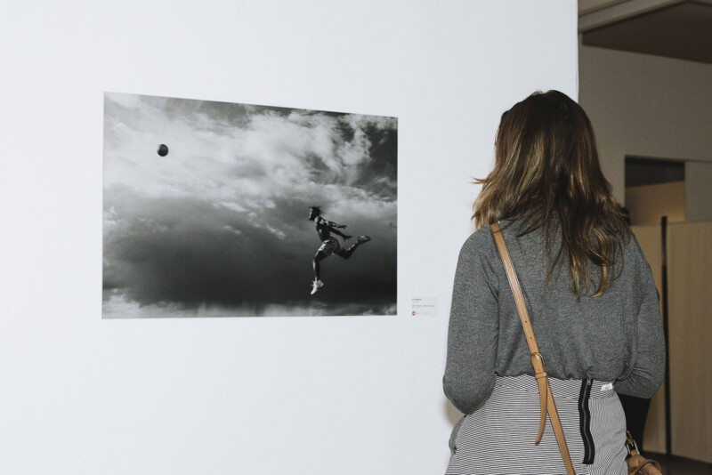 A person with long hair, seen from behind, observes a black-and-white photograph of a person jumping to kick a soccer ball against a cloudy sky, displayed on a white gallery wall.
