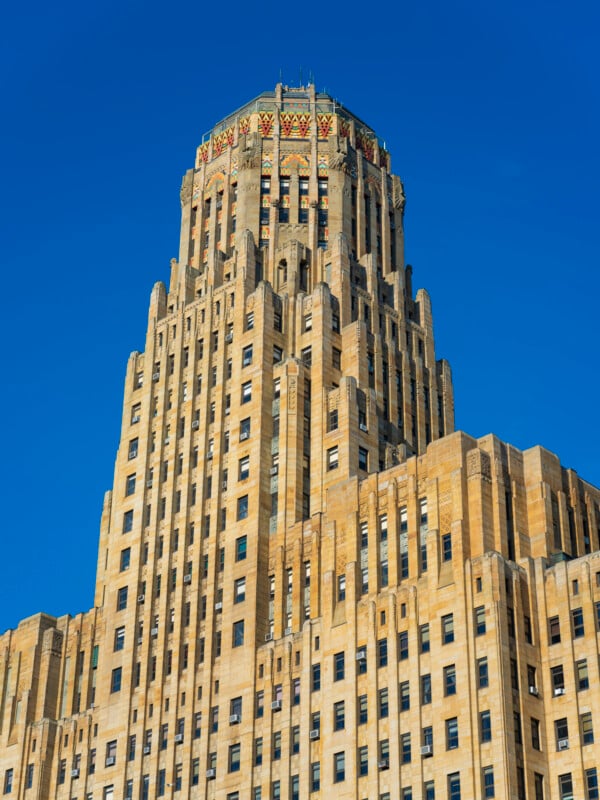 Art Deco-style skyscraper with tan stone exterior, vertical lines, and a colorful ornate crown on top, set against a clear blue sky.