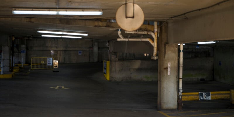 Dimly lit underground parking garage with concrete pillars and yellow barriers, showing "One Way" and "No Parking" signs, pipes on the ceiling, and directional arrows painted on the ground.