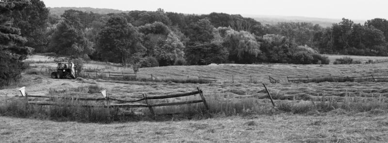 A black and white photo of a rural landscape with rolling fields, tall trees, and a wooden fence. A tractor is parked on the left side, and hay is spread across the ground in the foreground.