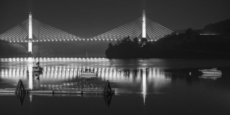 Black and white photo of a cable-stayed bridge illuminated at night, reflecting on calm water with several boats anchored in the foreground and a tree-covered shoreline in the background.