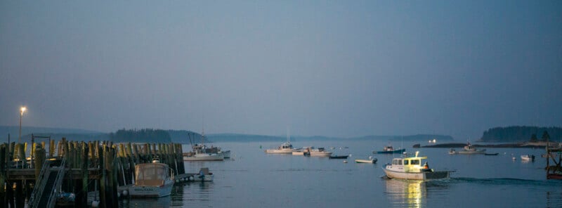 Boats float on calm water at dusk near a wooden dock, with distant tree-lined islands and a hazy sky in the background; one boat is illuminated and moving in the foreground.