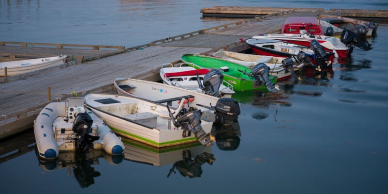 Several small motorboats and dinghies are moored alongside a wooden dock on calm water, with reflections visible and various boats in white, green, and red colors.