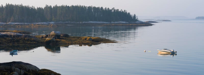 Calm coastal scene with rocky shores, evergreen trees, and two small boats floating on still water under a hazy sky.