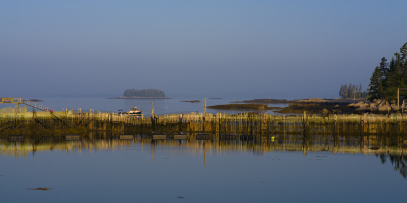 A calm coastal scene at sunrise shows a wooden pier, lobster traps, and a small boat on still water, with small islands and trees in the distance under a clear blue sky.