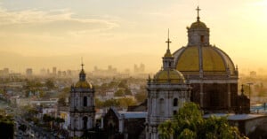 A scenic view of Mexico City at sunset, featuring the domes of a historic church in the foreground with modern city buildings and a hazy sky in the background.