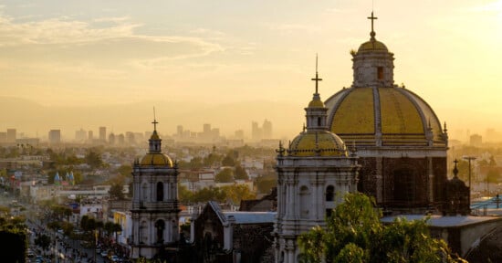 A scenic view of Mexico City at sunset, featuring the domes of a historic church in the foreground with modern city buildings and a hazy sky in the background.