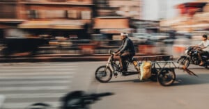 A man rides a motorbike with an attached cart down a busy street, motion blur showing speed; another motorcyclist follows, with buildings and pedestrians in the blurred background.