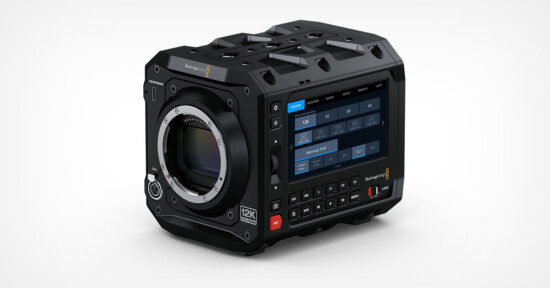 Close-up of a black, rectangular 12K digital cinema camera with a large lens mount on the front and a touchscreen display showing settings on the side. The camera has various buttons and vents on its sturdy body.