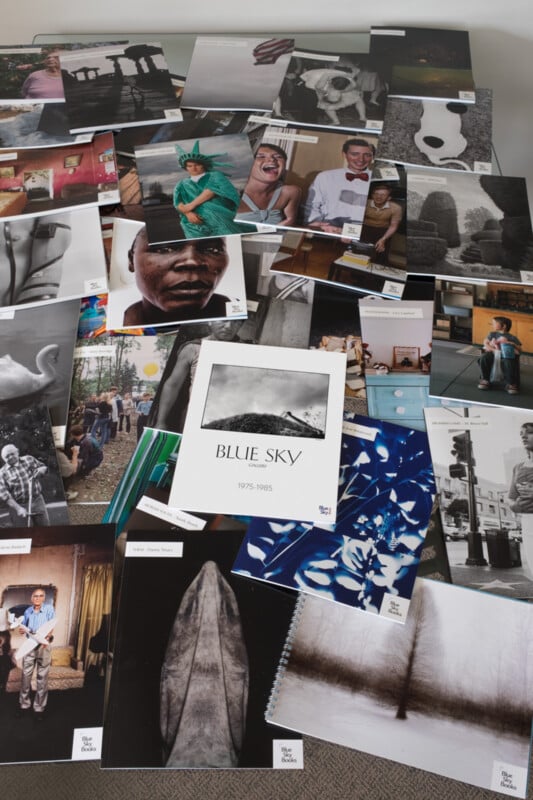 A large assortment of colorful and black-and-white photographs is spread out on a flat surface, including portraits, landscapes, and abstract images. In the center is a book titled "Blue Sky 1975-1985.
