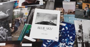 A variety of photography exhibition flyers and postcards are spread out, with a central catalog titled "Blue Sky Gallery 1975-1985" featuring a black-and-white photo of a hill with a house at the top.