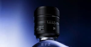 A black camera lens with "24" marked on it stands upright on a reflective, curved surface against a gradient blue and black background. The lens has focus and zoom rings, a switch, and engraved text.