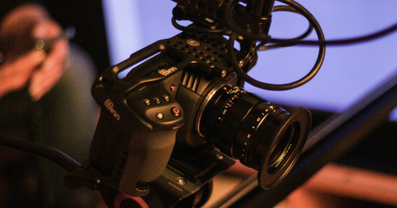 A close-up of a professional video camera with cables attached, set up on a tripod against a blurred background.