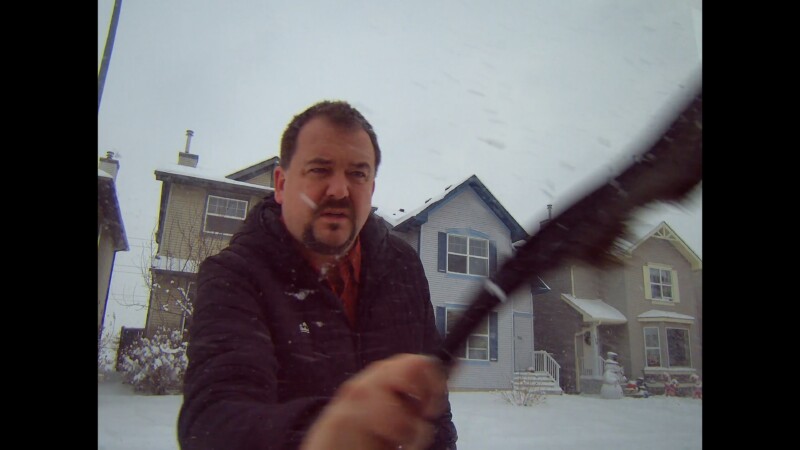A man wearing a dark jacket clears snow from a camera lens with a brush outside, with snow falling and several houses in the background on a winter day.