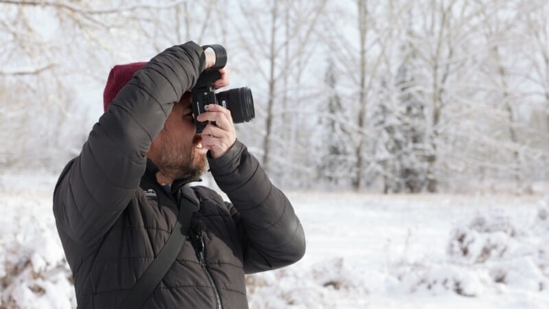 A person wearing a dark winter jacket and red beanie is taking a photo with a camera outdoors in a snowy landscape, with leafless trees in the background.