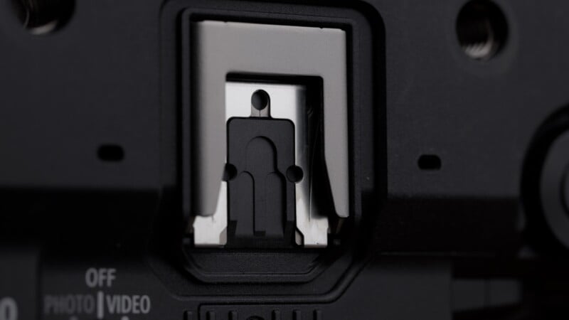 Close-up of a camera hot shoe mount with a small plastic cover in place, featuring an abstract outline of a person on the cover. The surrounding camera body is black with some control labels visible.