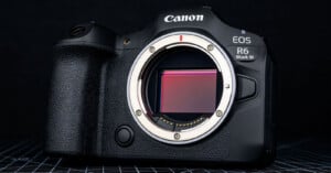 A Canon EOS R6 Mark III mirrorless camera body is shown without a lens, revealing its image sensor, on a dark, grid-patterned surface.