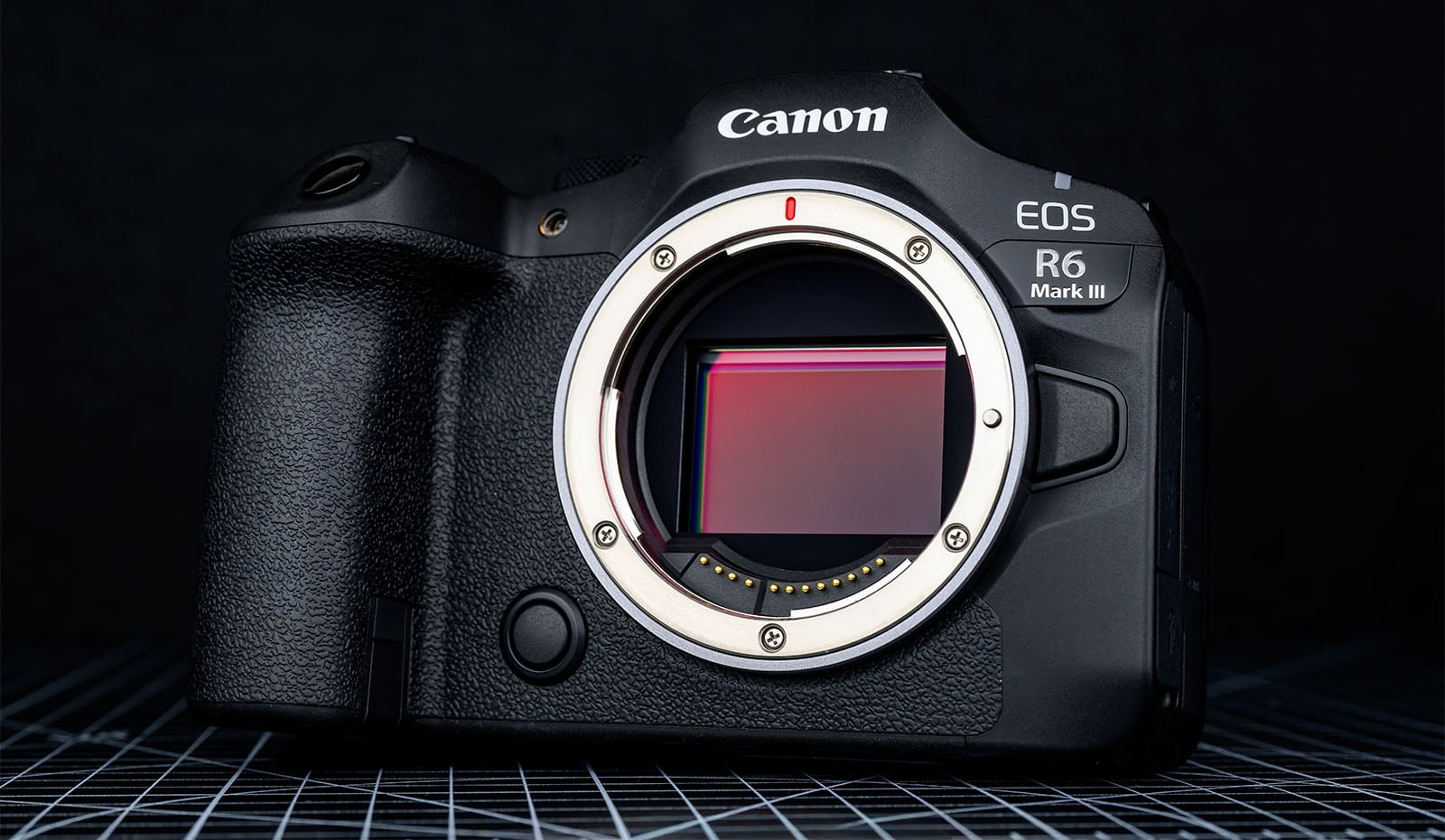 A Canon EOS R6 Mark III mirrorless camera body without a lens, showing the camera sensor, placed on a grid-patterned surface with a dark background.