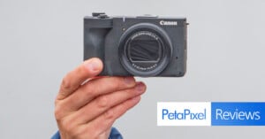 A hand holds a compact Canon camera in front of a plain background. The bottom right corner features the "PetaPixel Reviews" logo.