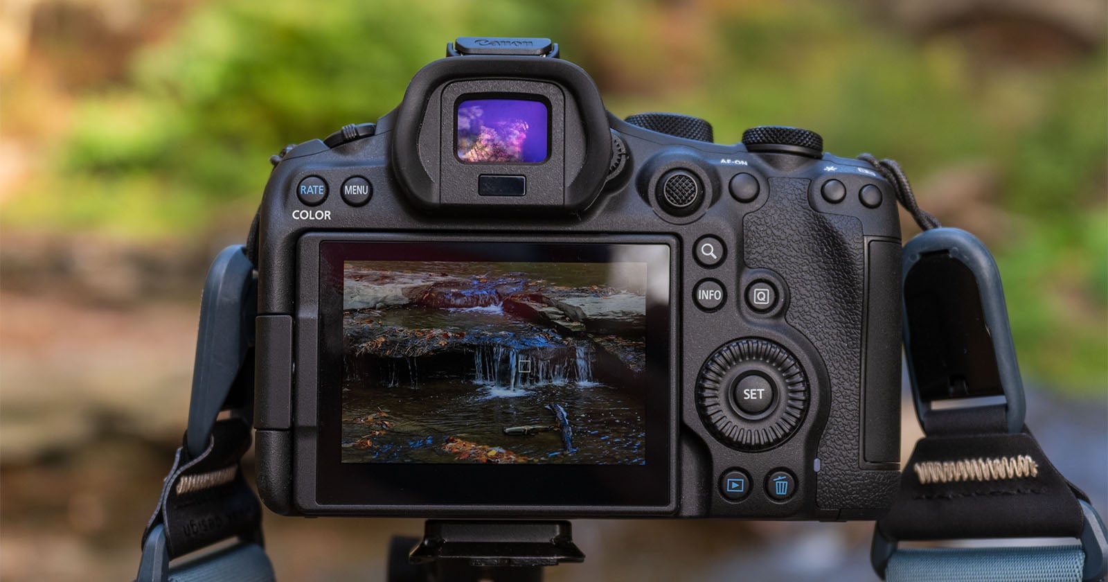 A digital camera on a tripod displays a close-up photo of a small waterfall on its screen; the blurred natural background suggests the camera is outdoors capturing a nature scene.