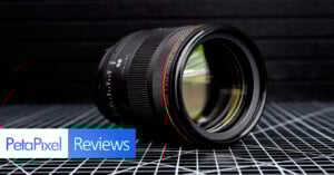 A camera lens rests on a black grid-patterned surface with a dark, lined background. The words "PetaPixel Reviews" appear in blue and white boxes at the bottom left corner.