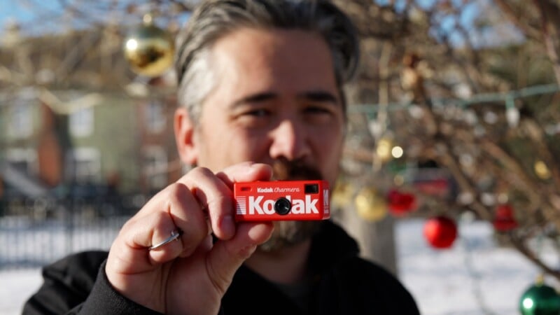 A person with gray hair holds a small Kodak film box close to the camera, while standing outdoors near trees decorated with colorful ornaments. The background is slightly blurred.
