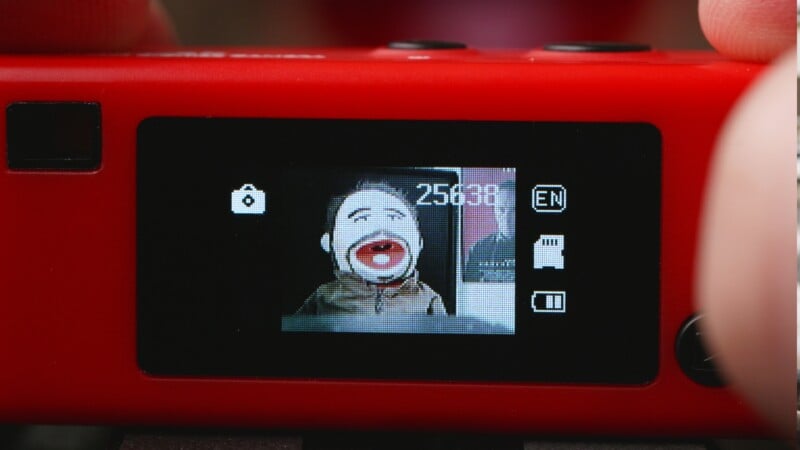 A close-up of a red digital camera screen displaying a photo preview of a puppet with brown hair and a wide open mouth. Camera settings and icons are visible around the preview image.