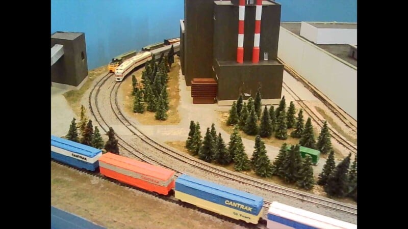 A model train set features a curved track with several boxcars, a locomotive, trees, and a large building with red-and-white smokestacks in the background, surrounded by scattered greenery.