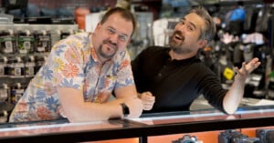 Two men stand behind a glass display counter in a camera store; one wears a floral shirt and leans forward, the other in a black shirt gestures playfully with a smile. Camera gear and accessories are visible in the background.