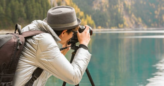 A person wearing a gray hat and jacket takes a photo with a camera on a tripod beside a calm lake, with trees and mountains in the background.