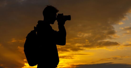 Silhouette of a person with a backpack taking a photo with a camera against a dramatic, golden sunset sky with scattered clouds.