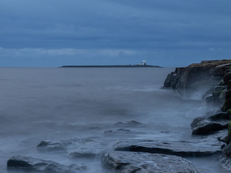 Rocky coastline with misty waves in the foreground, under a cloudy blue sky. In the distance, an island with a lighthouse and lit buildings is visible across the calm sea.
