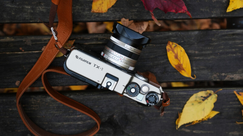 A Fujifilm TX-1 camera with a brown leather strap sits on a weathered wooden bench covered with scattered yellow and red autumn leaves.