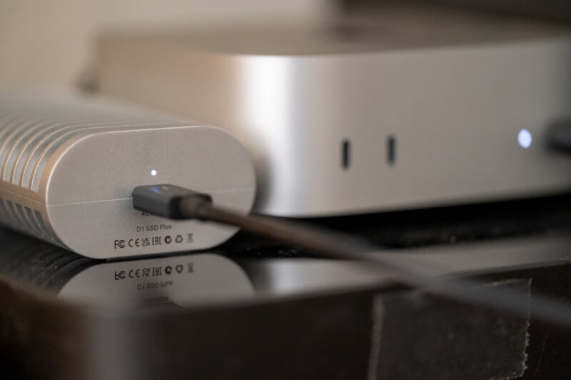 A close-up of an external SSD drive connected to a computer via a cable, with both devices resting on a dark reflective surface. The SSD has indicator lights and visible ports, and the computer is silver with a sleek design.