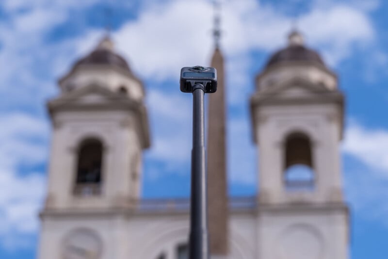 A 360-degree camera on a selfie stick is centered in the foreground, with two blurred church towers and a blue sky with clouds in the background.