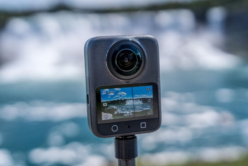 A 360-degree action camera on a tripod, with its small display screen showing a live preview of a scenic, blurred waterfall background.