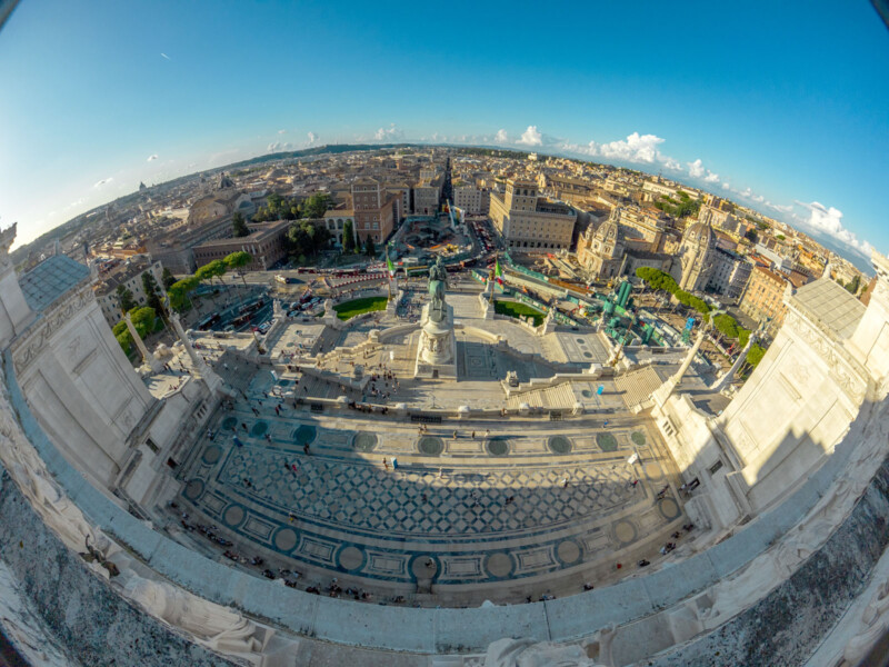 A wide-angle, fisheye view from atop a grand marble building overlooking Rome, Italy, capturing city rooftops, streets, and a large open plaza with intricate geometric patterns. Blue sky and distant clouds are visible.
