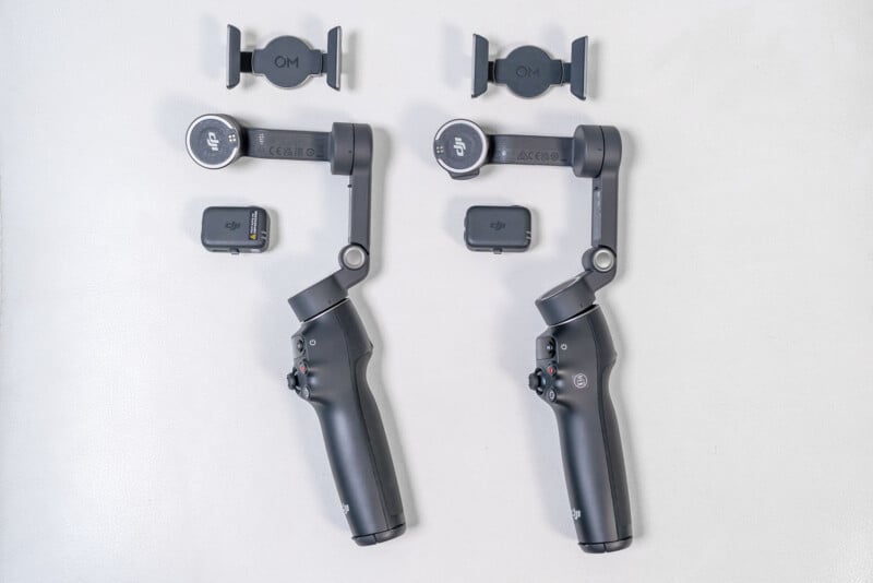 Two black handheld gimbals with detachable phone holders and small accessories are displayed side by side on a white surface. The items appear identical, with all components shown separately.