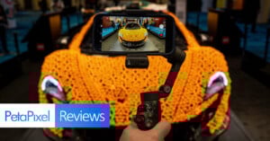 A person films a detailed LEGO car model using a smartphone mounted on a handheld gimbal. The LEGO car is bright orange and the setup suggests a tech or gadget review setting. "PetaPixel Reviews" is overlaid in the corner.