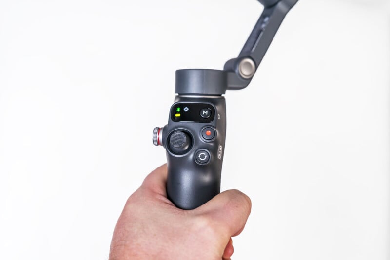 A hand holding a black camera gimbal stabilizer with control buttons and a small display, against a plain white background.